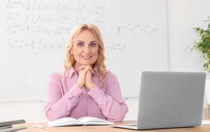 Happy teacher sitting near laptop at desk in classroom