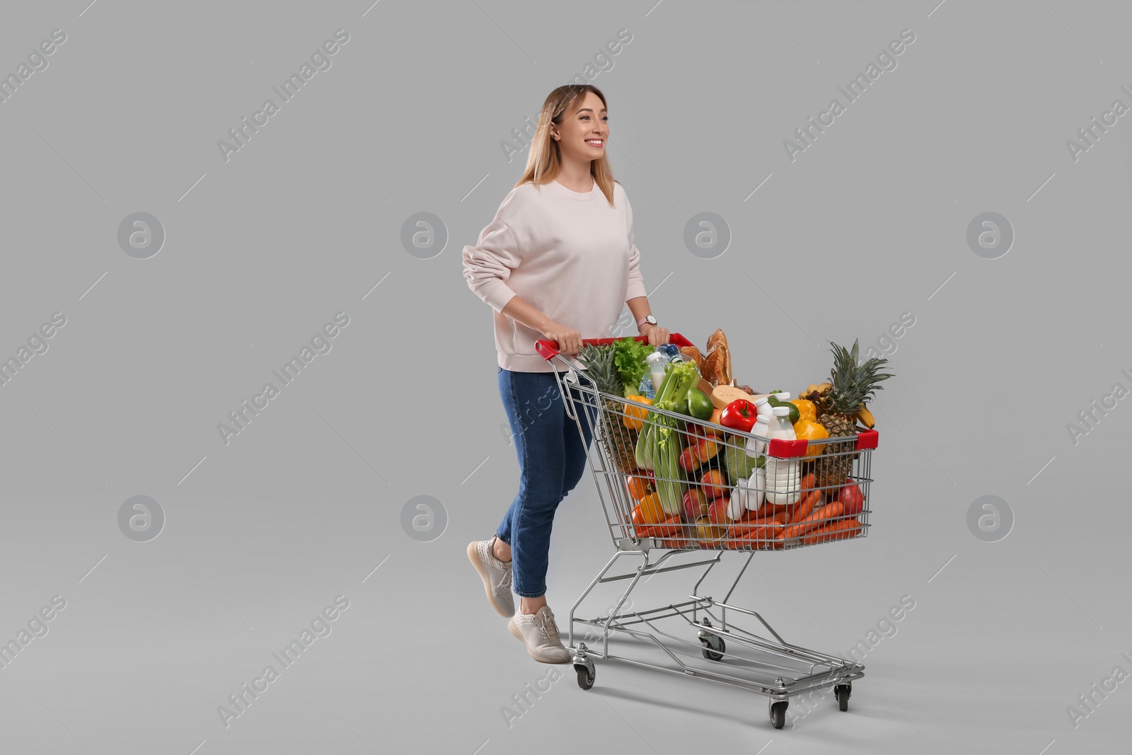 Photo of Young woman with shopping cart full of groceries on grey background