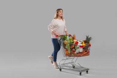 Young woman with shopping cart full of groceries on grey background