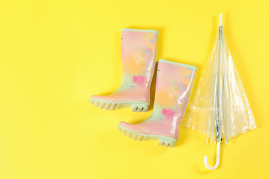 Photo of Transparent umbrella and colorful rubber boots on light yellow background, flat lay