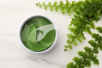Jar of under eye patches with spoon and green fern on white marble table, flat lay. Cosmetic product