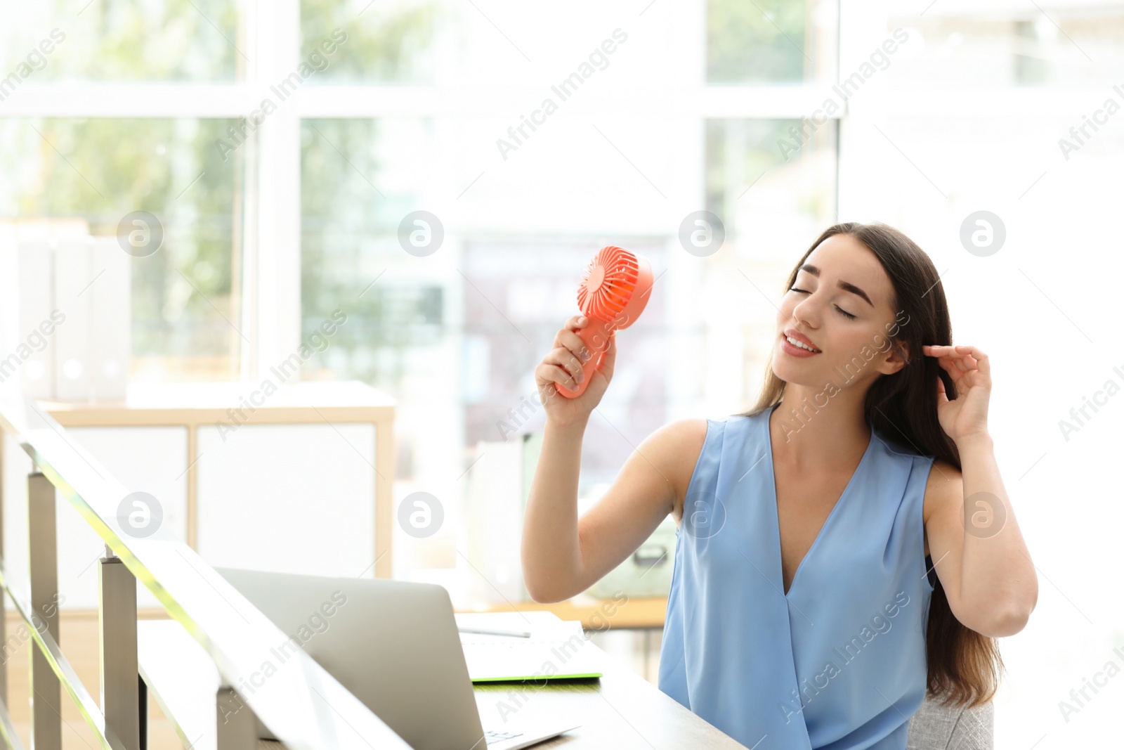 Photo of Young woman enjoying air flow from portable fan at workplace, space for text. Summer heat