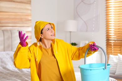 Photo of Young woman in raincoat collecting leaking water from ceiling at home. Time to call roof repair service