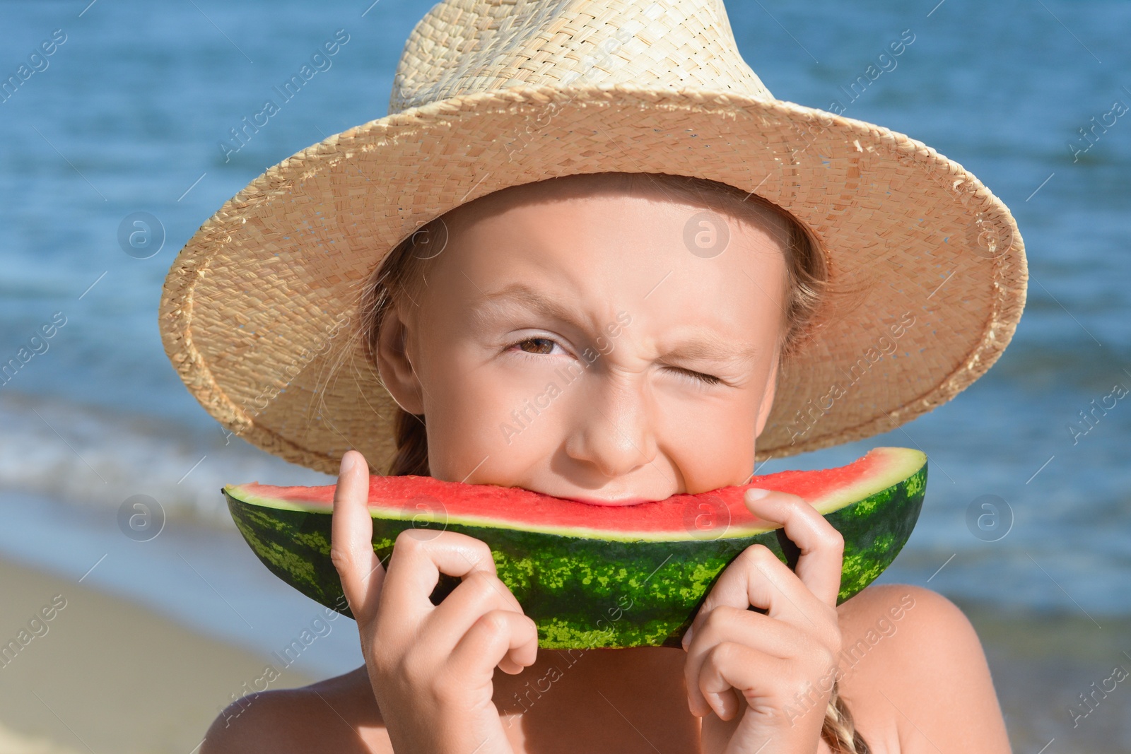 Photo of Cute little girl in straw hat eating juicy watermelon on beach, closeup