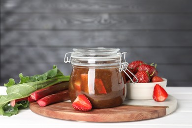 Photo of Jar of tasty rhubarb jam, fresh stems and strawberries on white table. Space for text