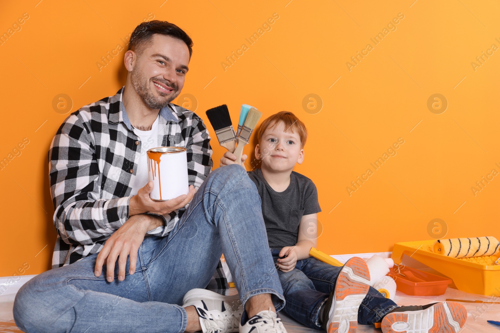 Photo of Father and son holding paint with brushes near orange wall, space for text. Repair work