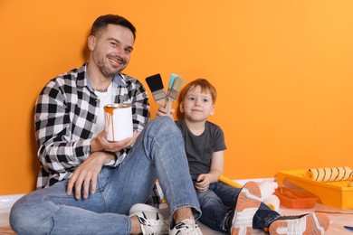 Photo of Father and son holding paint with brushes near orange wall, space for text. Repair work