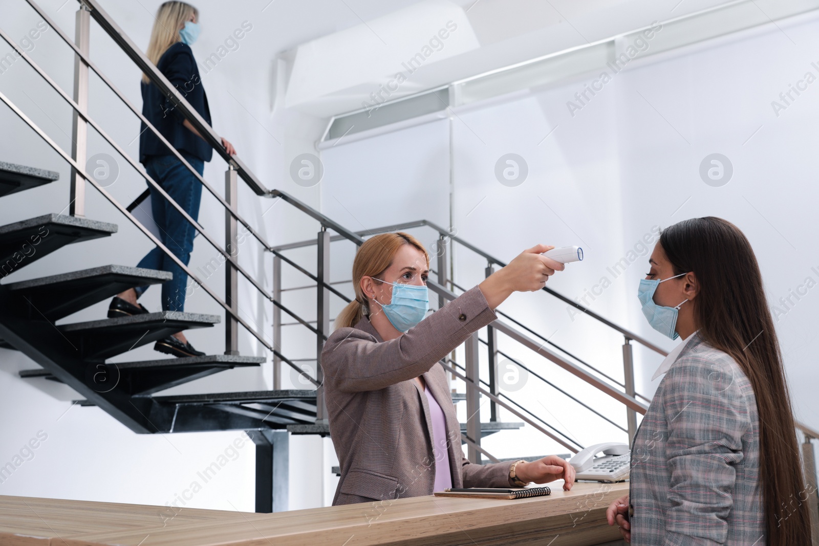 Photo of Woman in mask measuring temperature of employee with noncontact thermometer at office reception