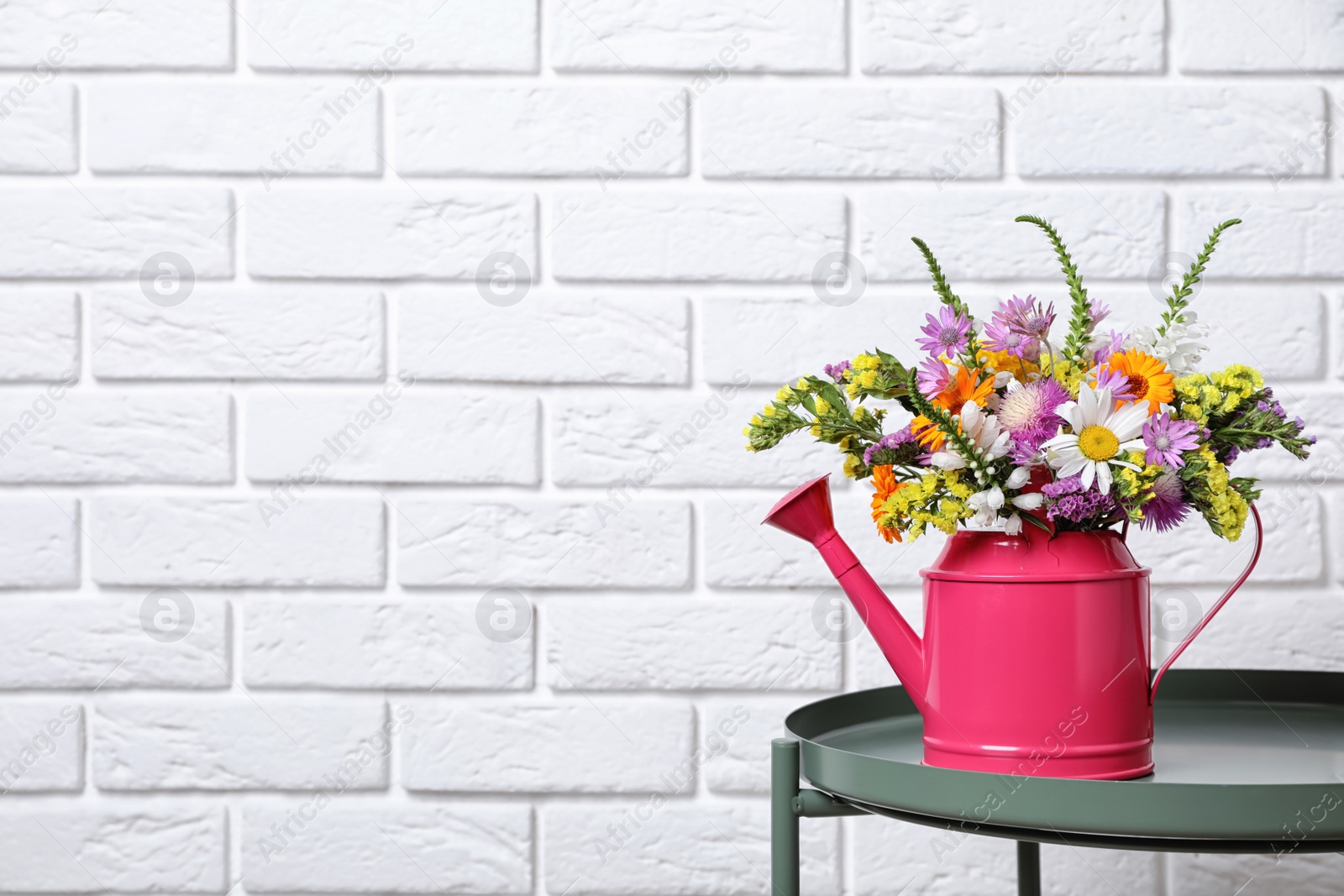 Photo of Watering can with beautiful wild flowers on table near brick wall