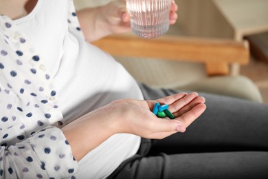Photo of Pregnant woman holding pile of pills and glass with water indoors, closeup