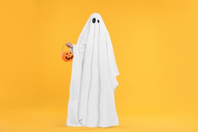 Photo of Woman in white ghost costume holding pumpkin bucket on yellow background. Halloween celebration