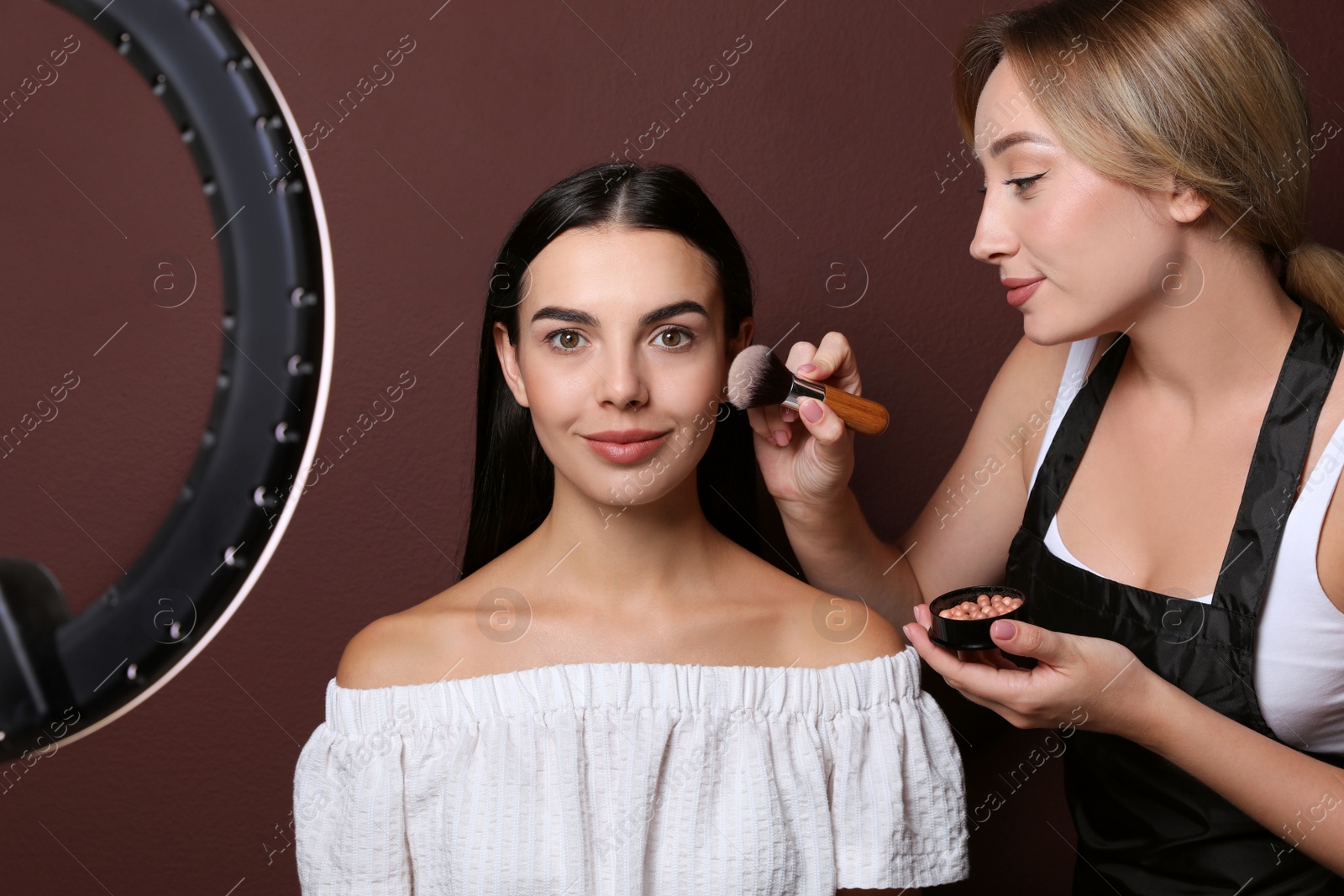 Photo of Professional makeup artist working with beautiful young woman against brown background. Using ring lamp