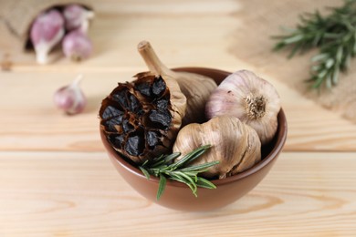 Photo of Bulbs of fermented black garlic and rosemary in bowl on wooden table