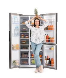 Photo of Young woman with pineapple on head near open refrigerator against white background