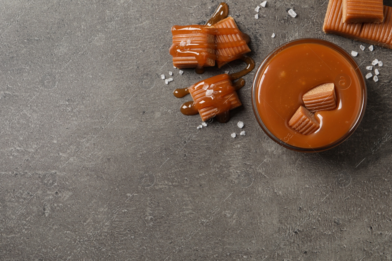 Photo of Tasty salted caramel with candies in glass bowl on grey table, flat lay. Space for text