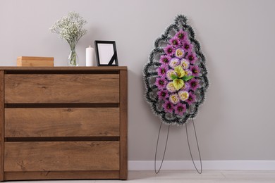 Photo of Wreath of plastic flowers and frame with black ribbon, burning candle on commode in room. Funeral attributes