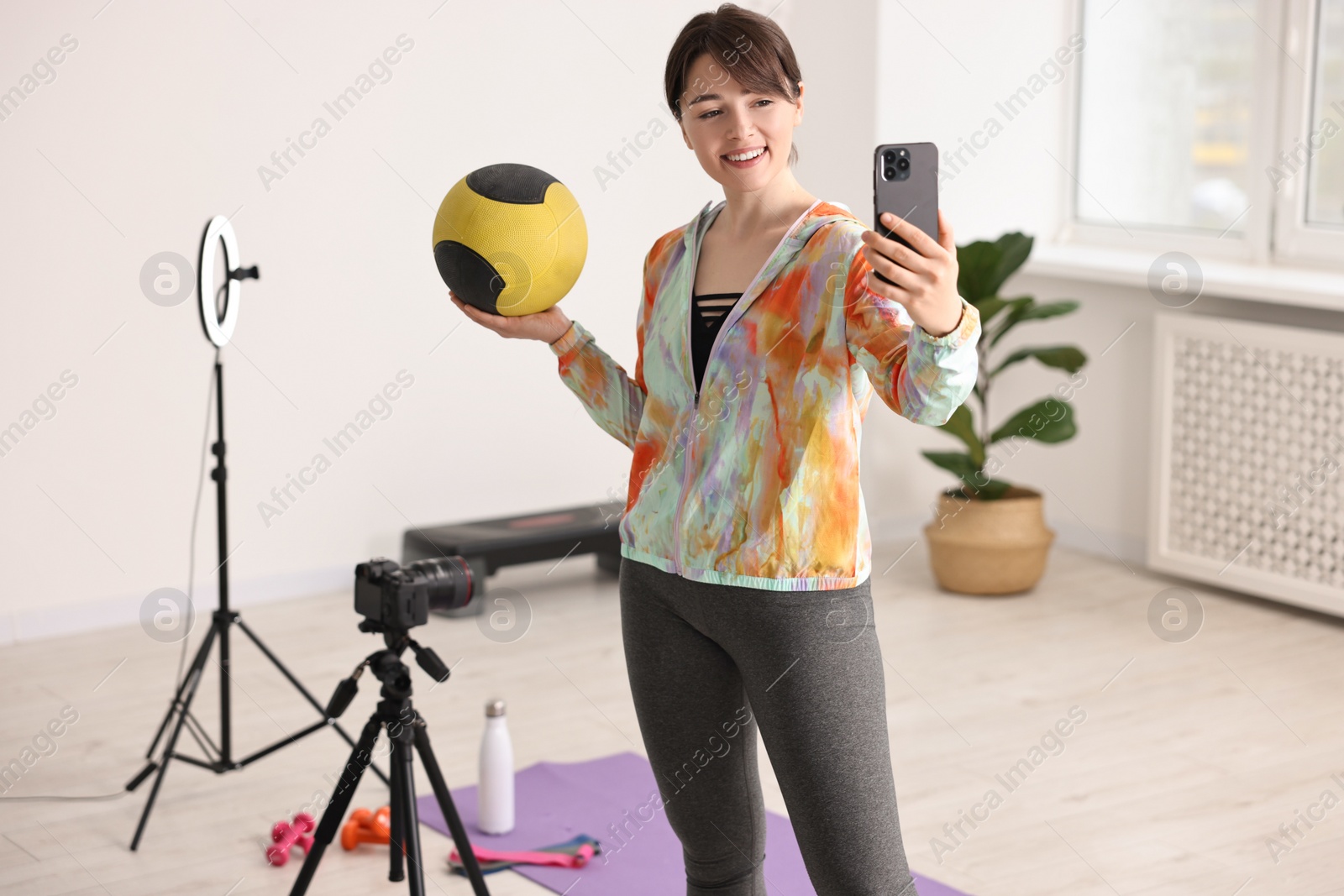 Photo of Happy sports blogger holding medicine ball while streaming online fitness lesson with smartphone at home