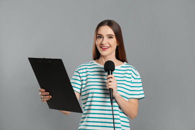 Young female journalist with microphone and clipboard on grey background