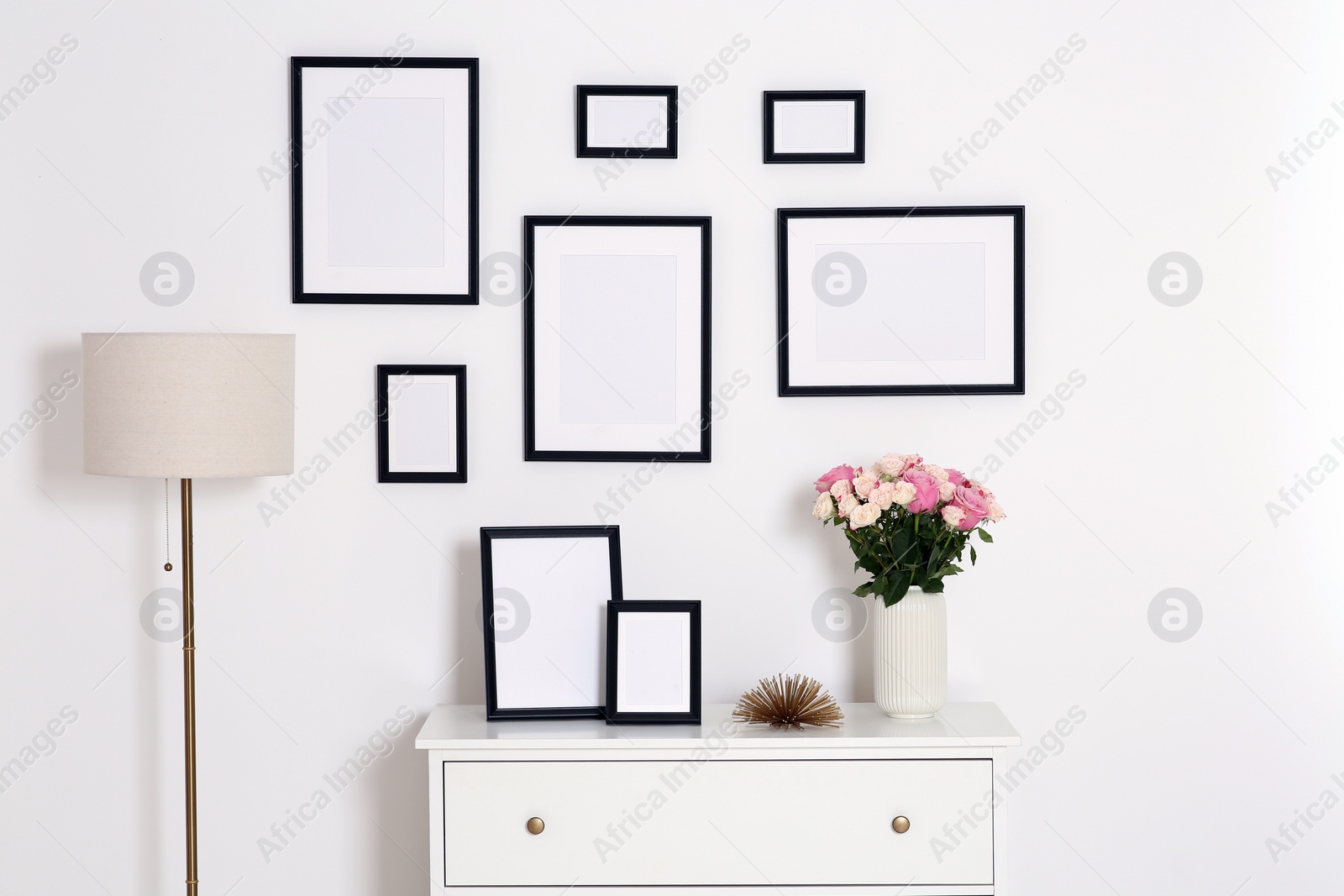 Photo of Empty frames hanging on white wall and chest of drawers with flowers indoors