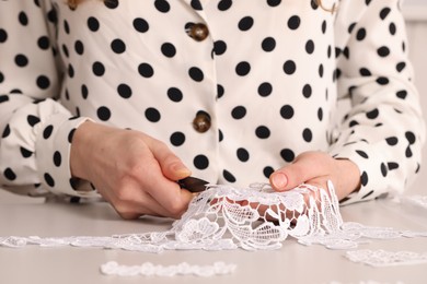 Photo of Dressmaker cutting beautiful white lace at table in atelier, closeup