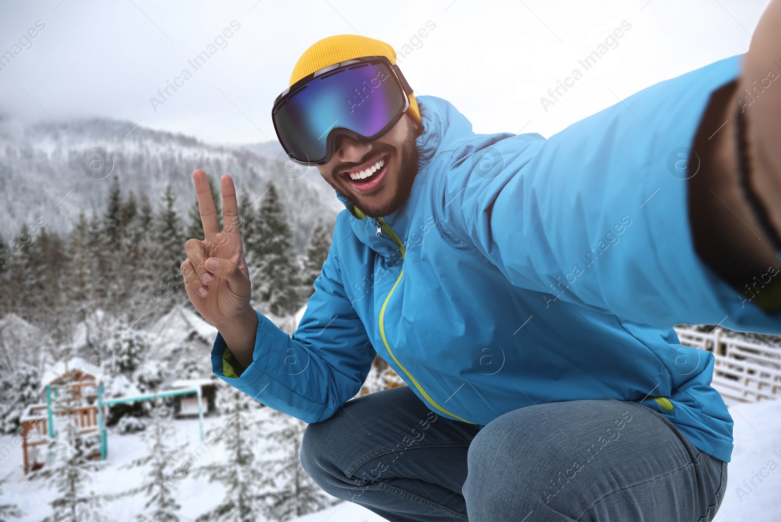 Image of Smiling young man in ski goggles taking selfie and showing peace sign in mountains