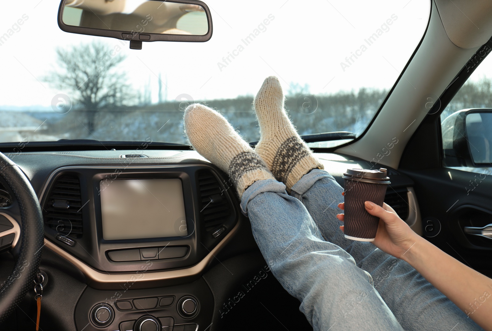 Photo of Young woman in warm socks holding her legs on car dashboard and drinking coffee. Cozy atmosphere