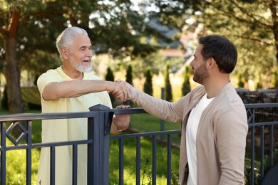 Photo of Friendly relationship with neighbours. Happy men shaking hands above fence outdoors