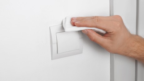 Photo of Man cleaning switch with disinfecting wipe on white wall indoors, closeup. Protective measures