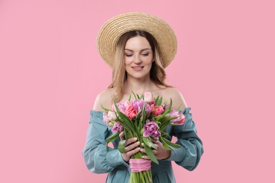 Happy young woman in straw hat holding bouquet of beautiful tulips on pink background