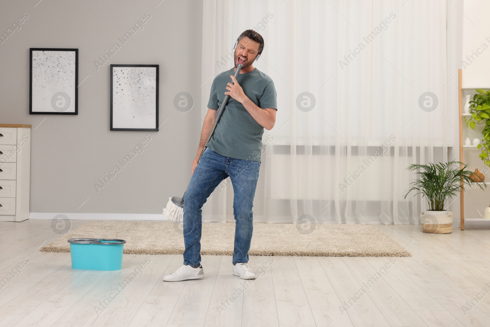 Photo of Happy man in headphones with mop singing while cleaning at home