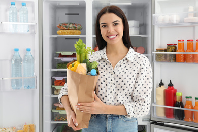 Photo of Young woman with bag of groceries near open refrigerator