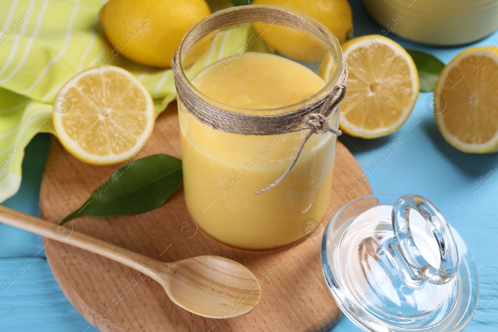 Photo of Delicious lemon curd in glass jar, fresh citrus fruits, green leaf and spoon on light blue wooden table
