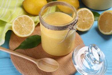 Delicious lemon curd in glass jar, fresh citrus fruits, green leaf and spoon on light blue wooden table