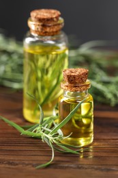 Photo of Bottles of essential oil and fresh tarragon leaves on wooden table, closeup