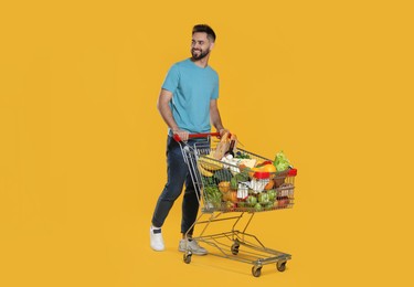 Photo of Happy man with shopping cart full of groceries on yellow background