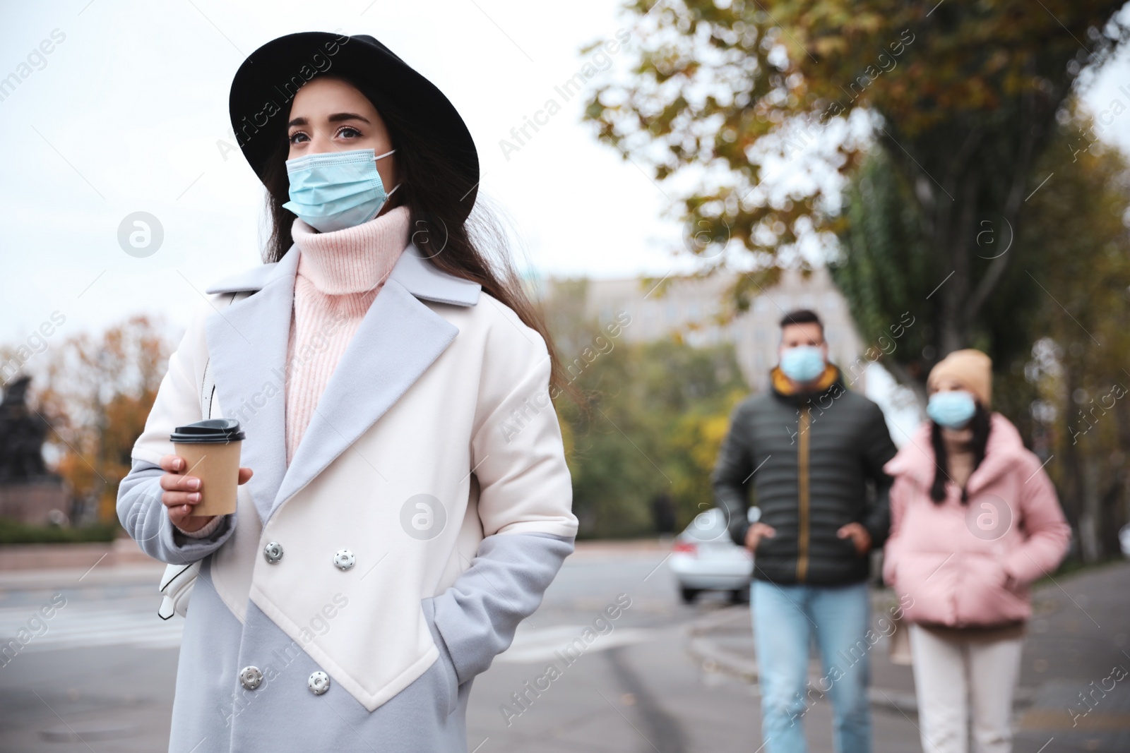 Photo of Young woman in medical face mask with cup of coffee walking outdoors. Personal protection during COVID-19 pandemic