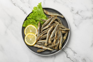 Plate with delicious fried anchovies, lemon and lettuce leaves on white marble table, top view
