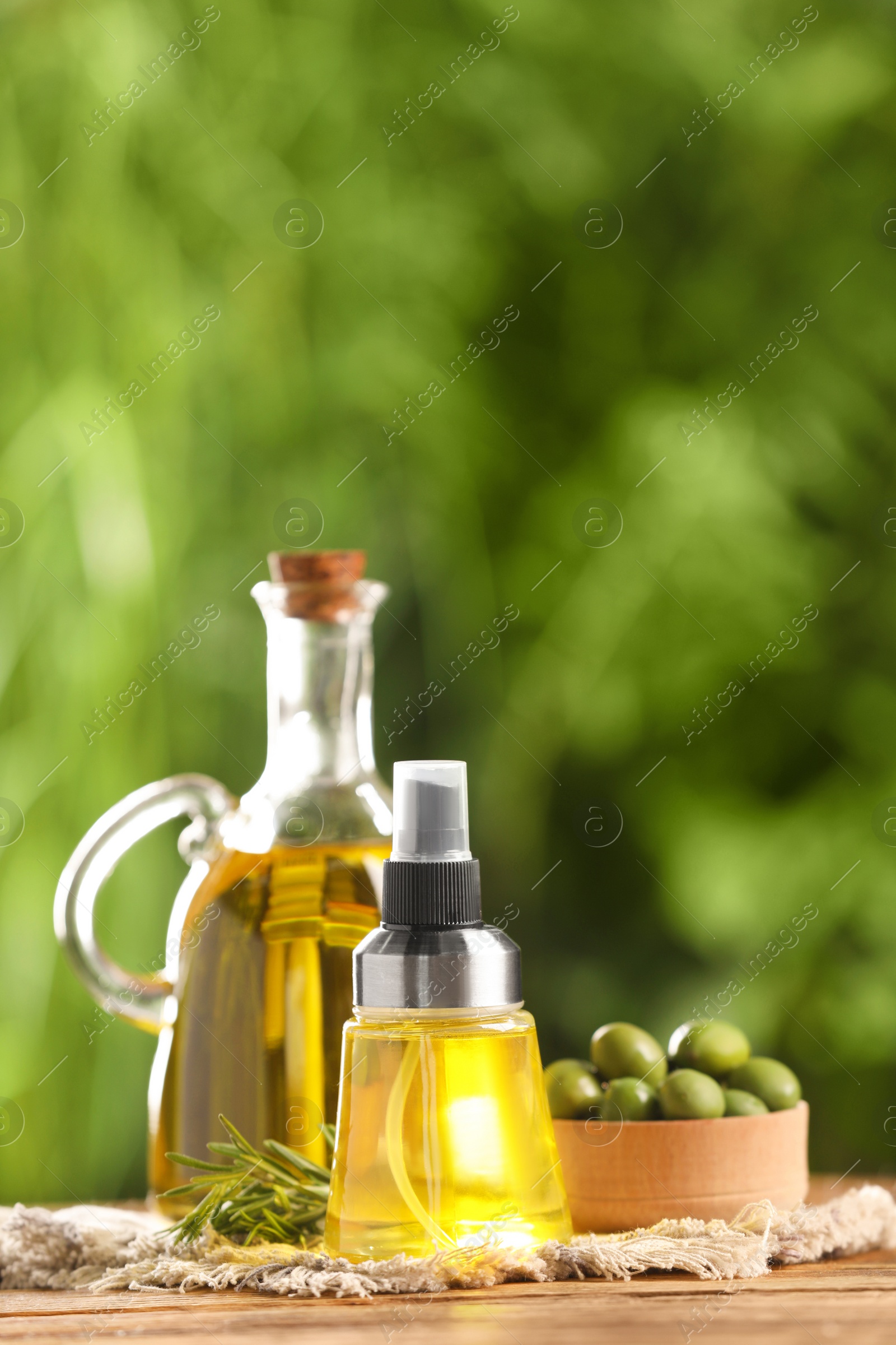 Photo of Bottles with cooking oil, olives and rosemary on wooden table against blurred background