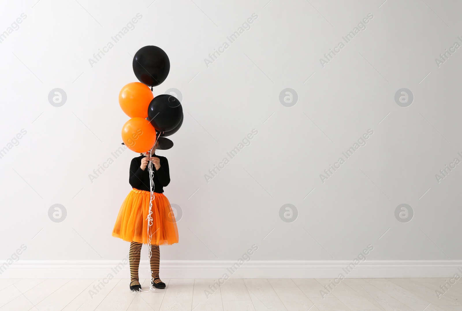 Photo of Cute little girl with balloons wearing Halloween costume near light wall. Space for text