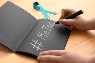 Photo of Woman writing hashtag MeToo in notebook near teal awareness ribbon at table, closeup. Stop sexual assault