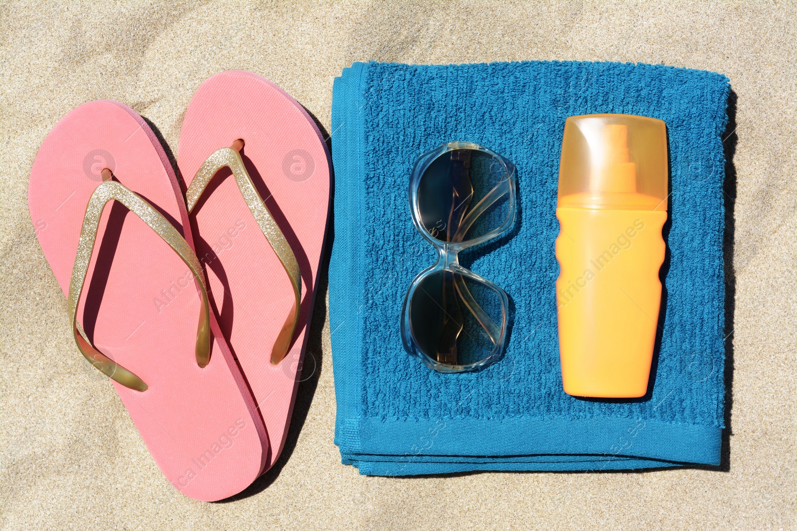 Photo of Folded soft blue beach towel with flip flops, sunglasses and bottle of sunscreen on sand, flat lay