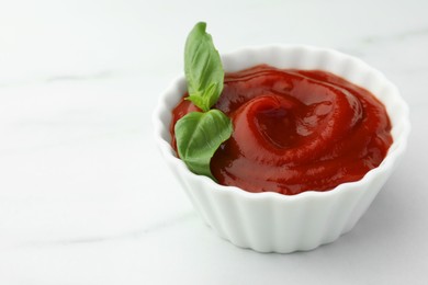 Photo of Bowl of tasty ketchup and basil on white marble table, closeup