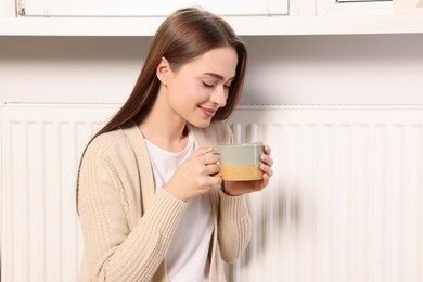 Woman holding cup with hot drink near heating radiator indoors