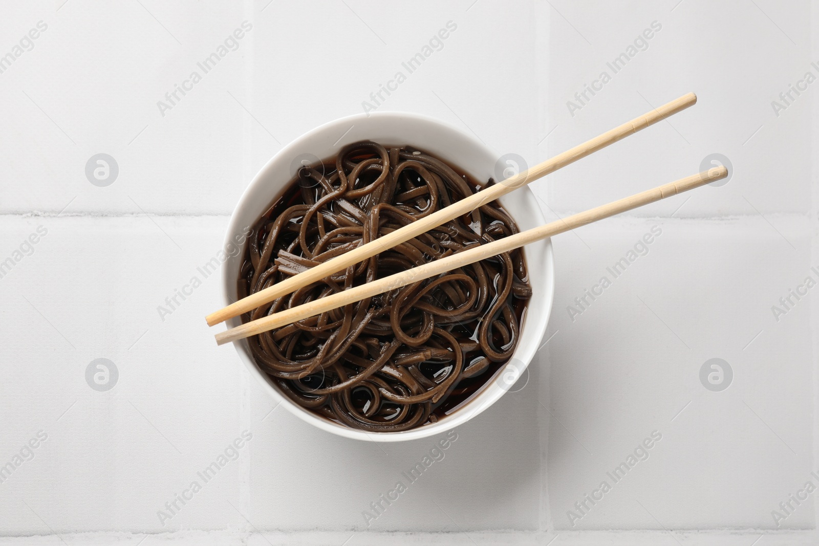 Photo of Tasty buckwheat noodles (soba) with sauce in bowl and chopsticks on white tiled table, top view