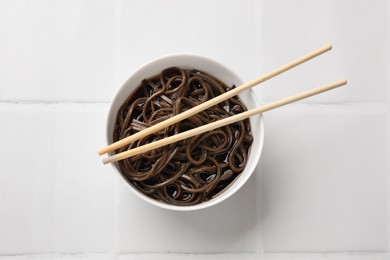 Photo of Tasty buckwheat noodles (soba) with sauce in bowl and chopsticks on white tiled table, top view