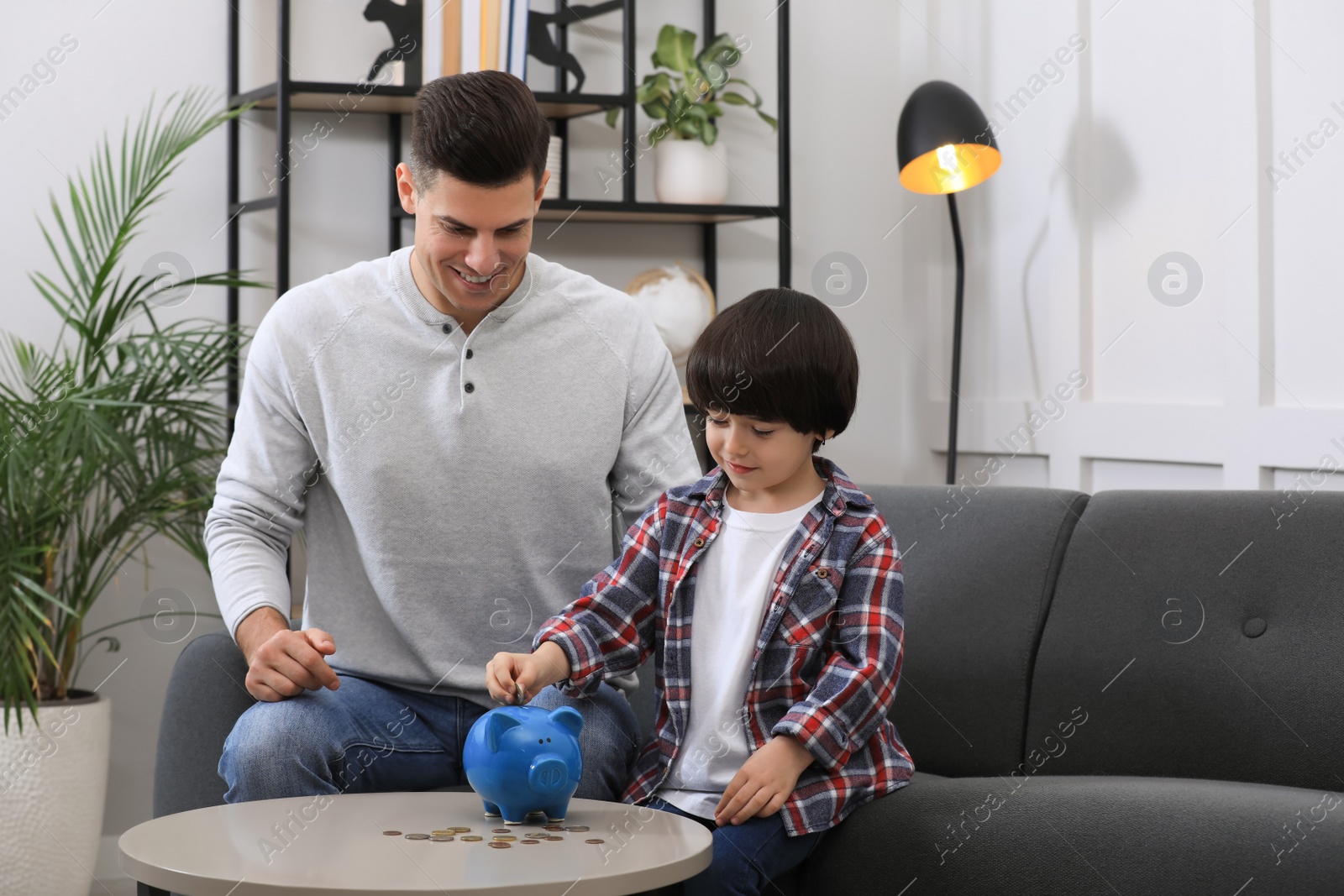 Photo of Little boy with his father putting coin into piggy bank at home