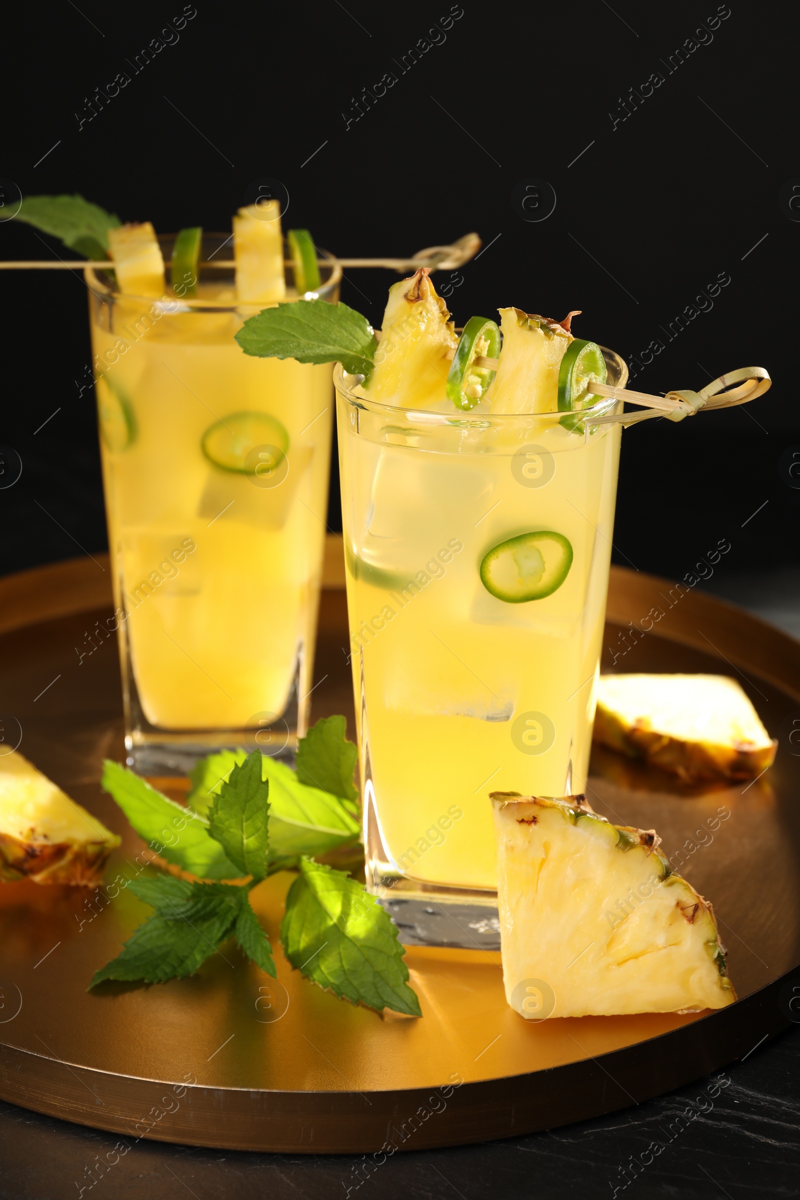 Photo of Glasses of tasty pineapple cocktail with sliced fruit, mint and chili pepper on black table