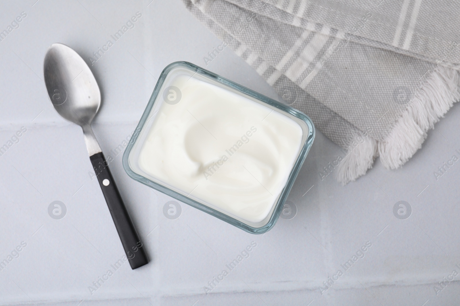 Photo of Delicious natural yogurt in glass bowl and spoon on white tiled table, top view