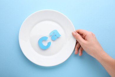 Photo of Woman holding plate with calcium symbol made of letters on light blue background, top view