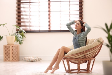 Young woman relaxing in papasan chair near window with blinds at home. Space for text